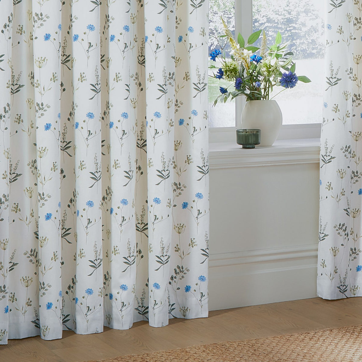 Meadow Floral cotton pencil pleat curtains in white with cornflower-blue and green wildflower print, pictured pleated at a window beside a vase of matching blue flowers.