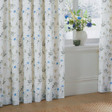 Meadow Floral cotton pencil pleat curtains in white with cornflower-blue and green wildflower print, pictured pleated at a window beside a vase of matching blue flowers.