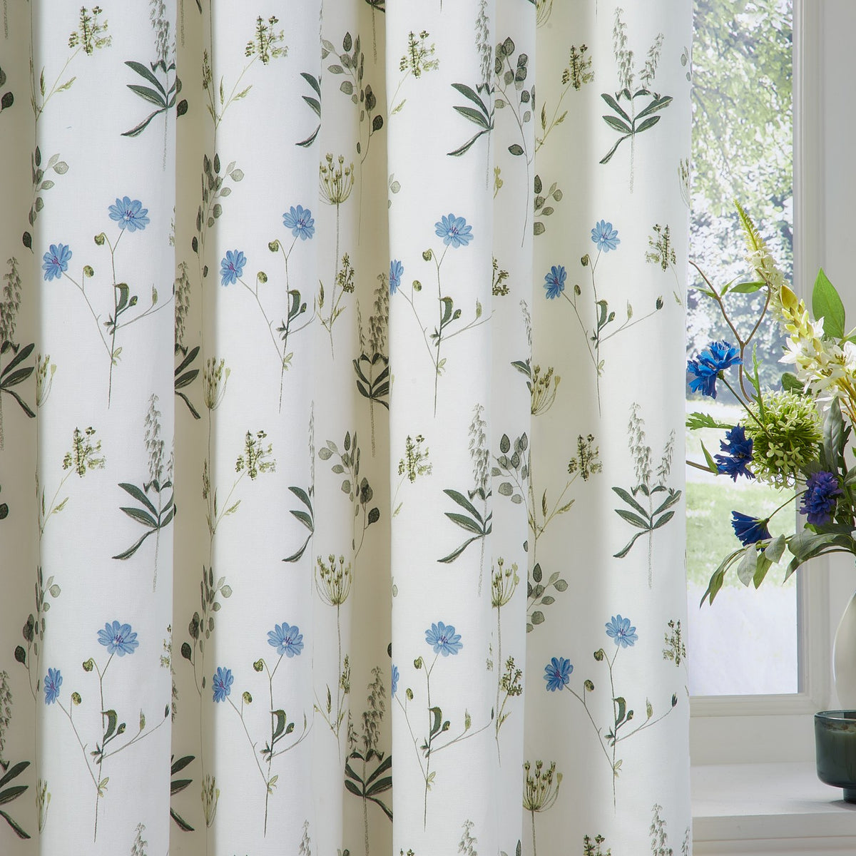 Meadow Floral cotton pencil pleat curtains in white with cornflower-blue blooms and green foliage, shown pleated by a window beside a vase of matching flowers.