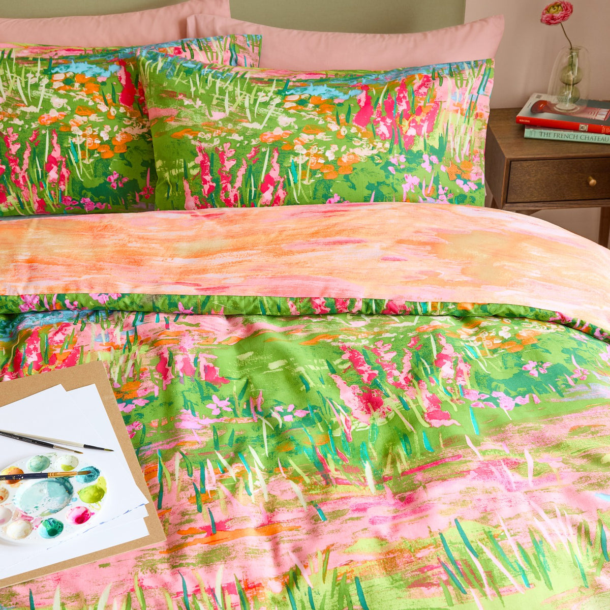 Meadow Escape reversible duvet cover set on a bed, showing green and pink wildflower meadow print pillowcases and a peach abstract reverse side, styled with a bedside table and paint palette.