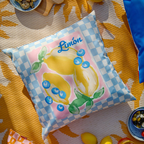 Square outdoor cushion with a pale blue and white checkerboard border framing a pink panel, hand-painted lemons, blueberries and leaves, with "Limón" lettered in blue.