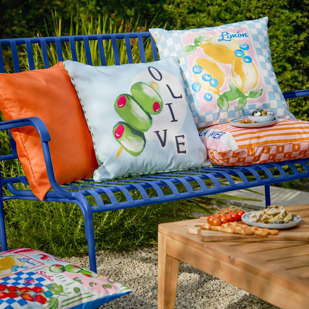 Mediterranean Limon reversible polyester cushion in sky blue with hand-painted lemon and blueberry print on pink, blue checkerboard border, shown on an outdoor bench with an olive-print cushion.
