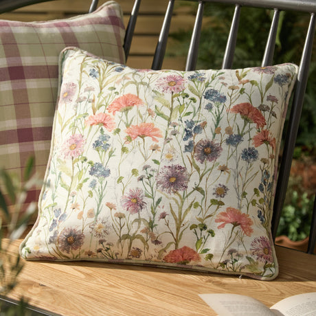 Medmerry Linen Cotton Cushion in Elderberry, 45 x 45cm square with hand-painted wildflower print in pink, orange and blue on cream linen, shown on an outdoor bench with a check cushion behind.