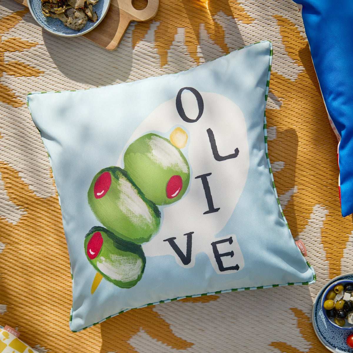 Mediterranean Olive Reversible Polyester Cushion in blue and green, showing a hand-painted olive skewer design with "OLIVE" text and striped piped trim, styled on an outdoor picnic setting.