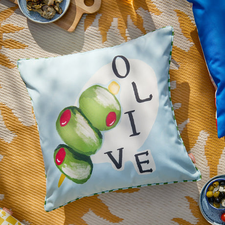 Mediterranean Olive Reversible Polyester Cushion in blue and green, showing a hand-painted olive skewer design with "OLIVE" text and striped piped trim, styled on an outdoor picnic setting.