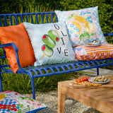 Olive/blue reversible polyester cushion with hand-painted olive design and green striped piped trim, on a blue garden bench styled with orange and citrus-print cushions.