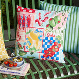 Mediterranean Picnic Abstract Polyester Cushion in multi patchwork print of sardines, olives, lemons, chillies and tomatoes on a green outdoor bench.