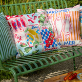 Mediterranean Picnic printed polyester cushions on a green garden bench, showing patchwork panels of sardines, olives, lemons, chillies and tomatoes in blue, red and pastel tones.