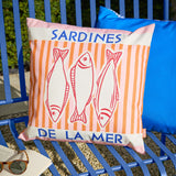 Square outdoor cushion in pink and orange stripes, printed with three red-line sardines and "SARDINES DE LA MER" text in blue, shown on a blue outdoor bench.
