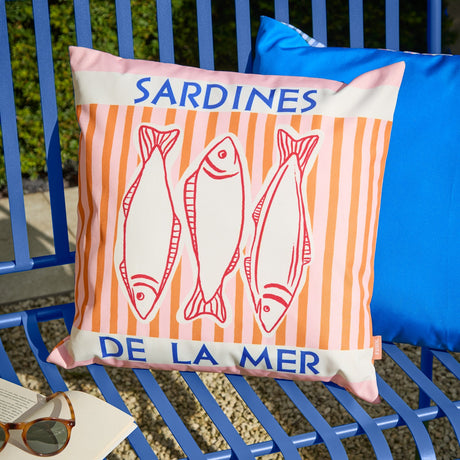 Square outdoor cushion in pink and orange stripes, printed with three red-line sardines and "SARDINES DE LA MER" text in blue, shown on a blue outdoor bench.
