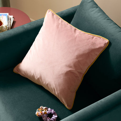 Meridian Plain Velvet Cushion in blush pink with gold piped edge, shown on a teal sofa in a living room setting.