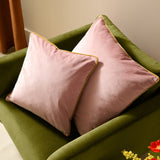 Two Meridian square velvet cushions in blush pink with gold piped edges, styled on an olive green sofa in a living room setting.