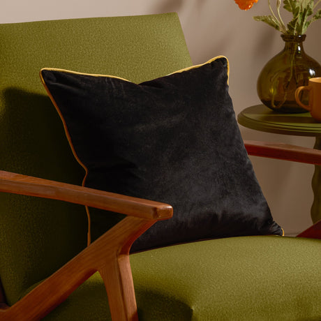 Meridian Plain Velvet Cushion in black with gold piped edging, shown on a green armchair beside a wooden side table.