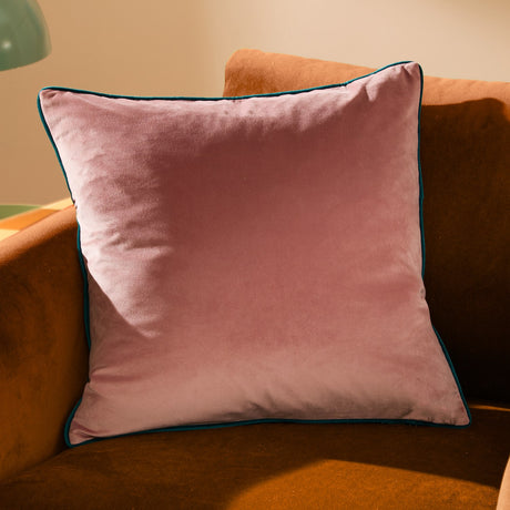 Meridian Plain Velvet Cushion in blush pink with teal piped edging, shown on a mustard velvet sofa in a sunlit living room setting.