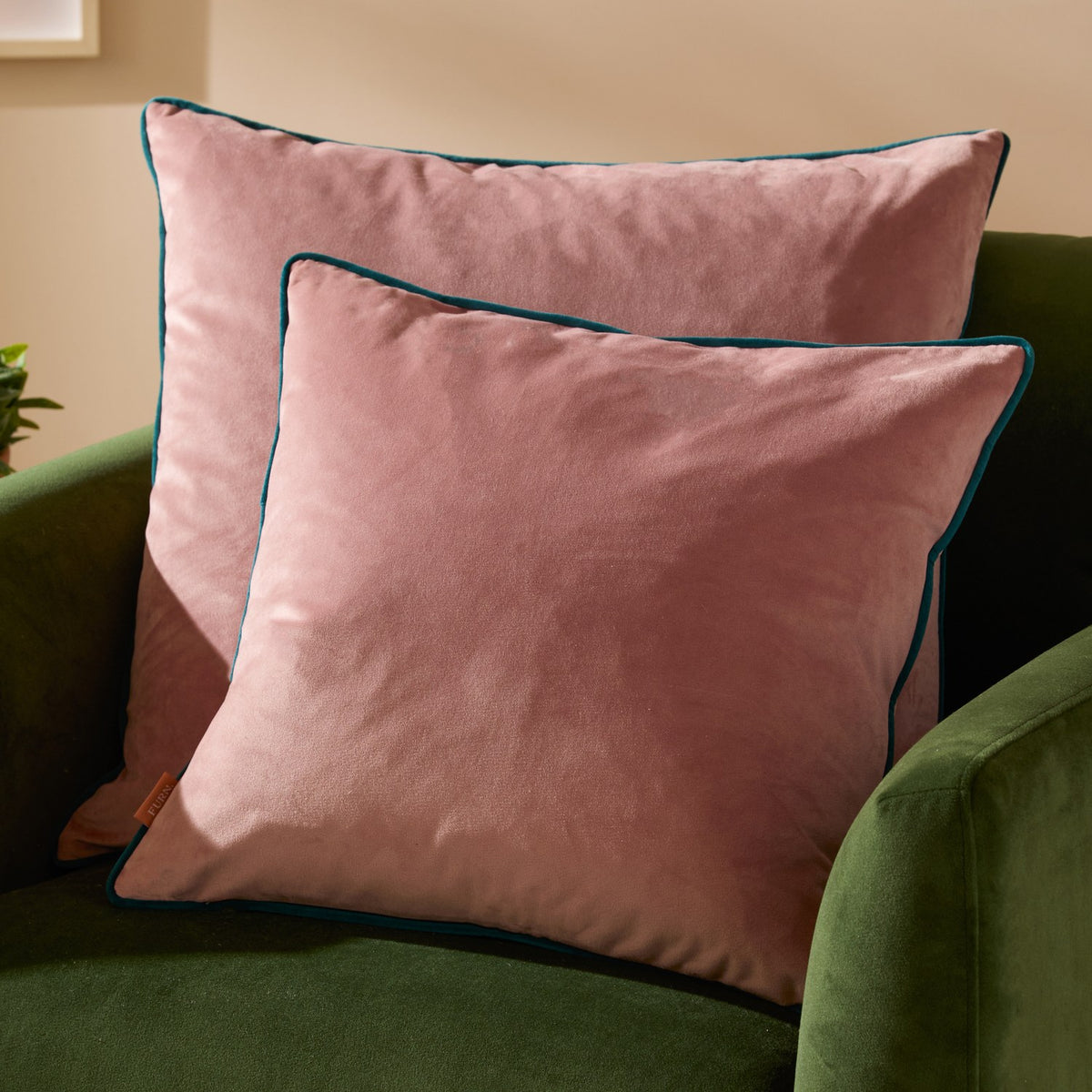 Two Meridian square velvet cushions in blush pink with contrasting teal piped edges, styled on a green velvet sofa.