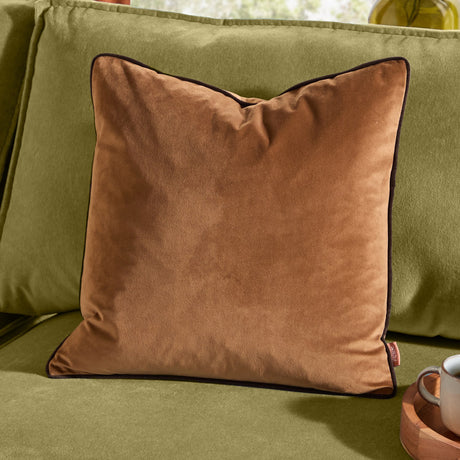 Meridian Plain Velvet Cushion in caramel with contrasting chocolate-brown piped edging, shown on a green sofa in a living room setting.