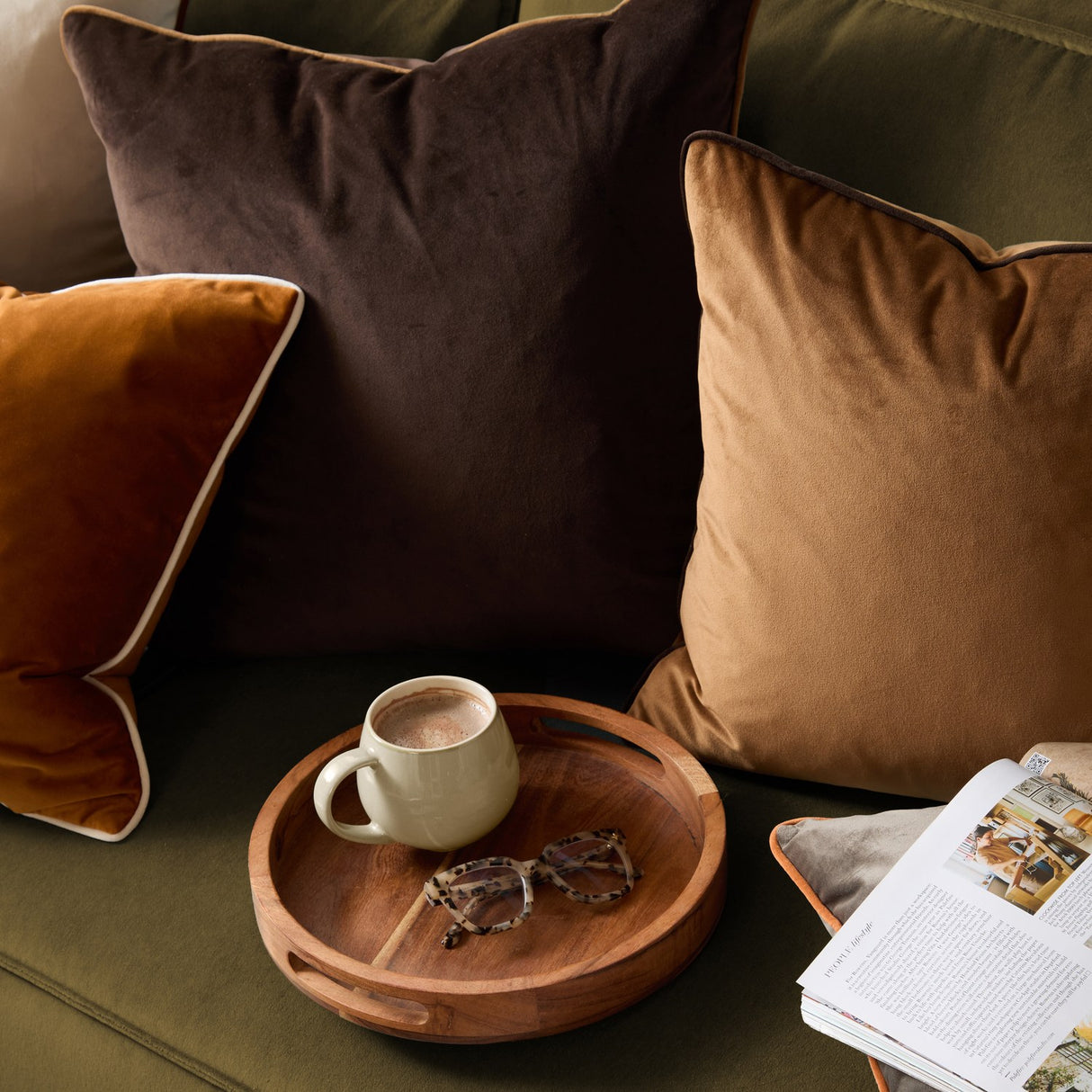Meridian Plain Velvet Cushion in caramel with chocolate-brown piped trim, shown on an olive-green sofa alongside coordinating brown and burnt-orange cushions, a wooden tray, mug, and magazine.