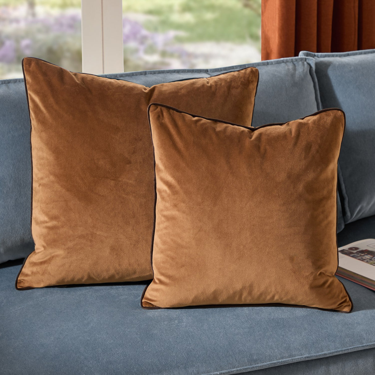Two Meridian plain velvet cushions in caramel with chocolate-brown piped edging, styled on a blue upholstered sofa near a window.