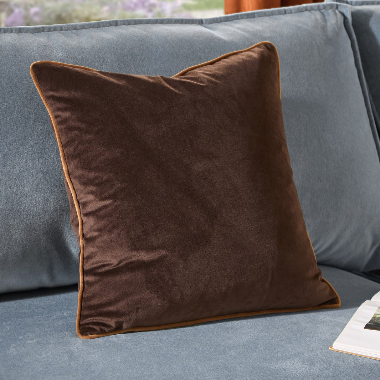 Meridian Plain Velvet Cushion in chocolate brown with contrasting caramel piped trim, shown on a light blue sofa.