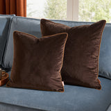 Two Meridian Plain Velvet Cushions in chocolate brown with caramel piped trim, styled on a blue sofa beside a window.