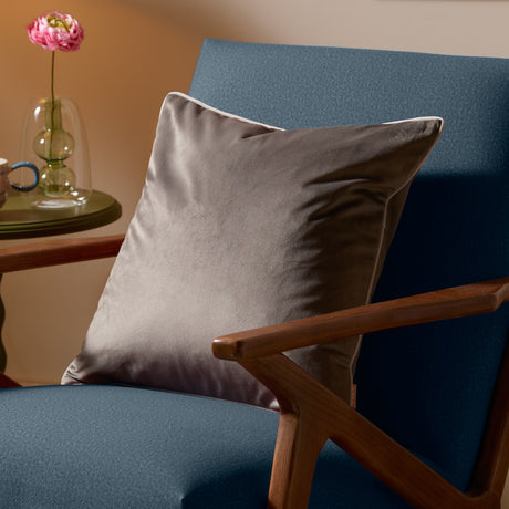 Meridian Plain Velvet Cushion in charcoal grey with dove piped edging, shown on a blue armchair beside a wooden side table with a vase and pink flower.