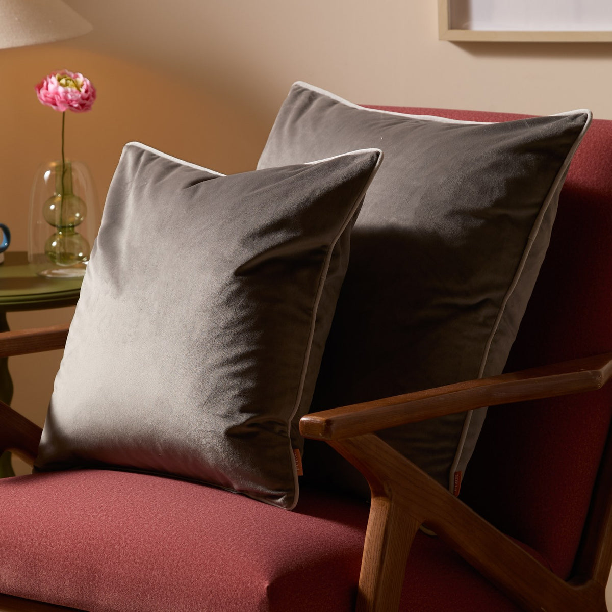 Two Meridian Plain Velvet Cushions in charcoal grey with dove piped edges, styled on a red upholstered armchair beside a wooden side table and pink flower.