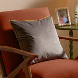 Meridian Plain Velvet Cushion in charcoal grey with moss green piped edge, styled on a rust upholstered armchair with wooden arms in a living room setting.