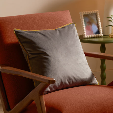 Meridian Plain Velvet Cushion in charcoal grey with moss green piped edge, styled on a rust upholstered armchair with wooden arms in a living room setting.