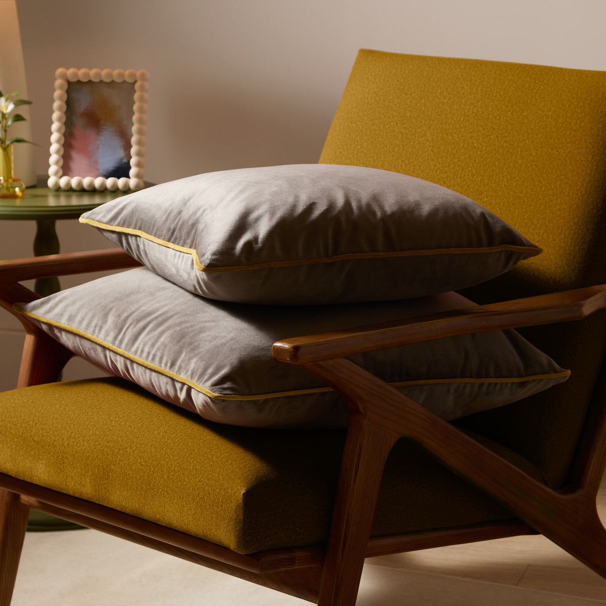 Two Meridian velvet cushions in charcoal grey with moss green piped edging, stacked on a mustard armchair with wooden frame.