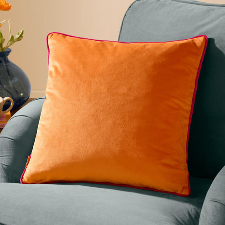 Meridian Plain Velvet Cushion in clementine orange with hot pink piped edging, shown on a light blue sofa beside a vase of orange flowers.