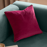 Meridian Plain Velvet Cushion in cranberry pink with mocha brown piped edge, square velvet design shown plumped on a teal sofa.