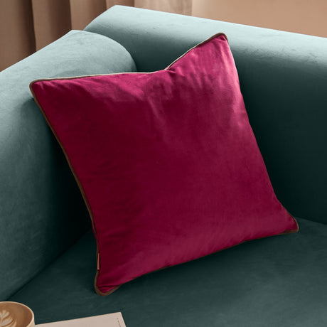Meridian Plain Velvet Cushion in cranberry pink with mocha brown piped edge, square velvet design shown plumped on a teal sofa.