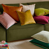 Meridian Plain Velvet Cushion in cranberry pink with mocha piping, shown among mixed velvet cushions in orange, mustard, pink, green and cream on an olive green sofa.