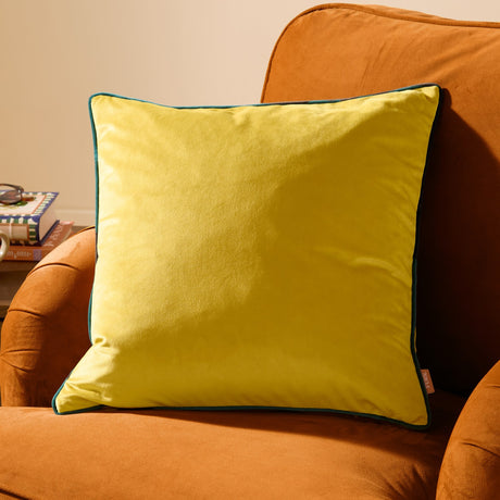 Meridian Plain Velvet Cushion in Cylon yellow with teal piped edging, shown resting against an orange armchair.