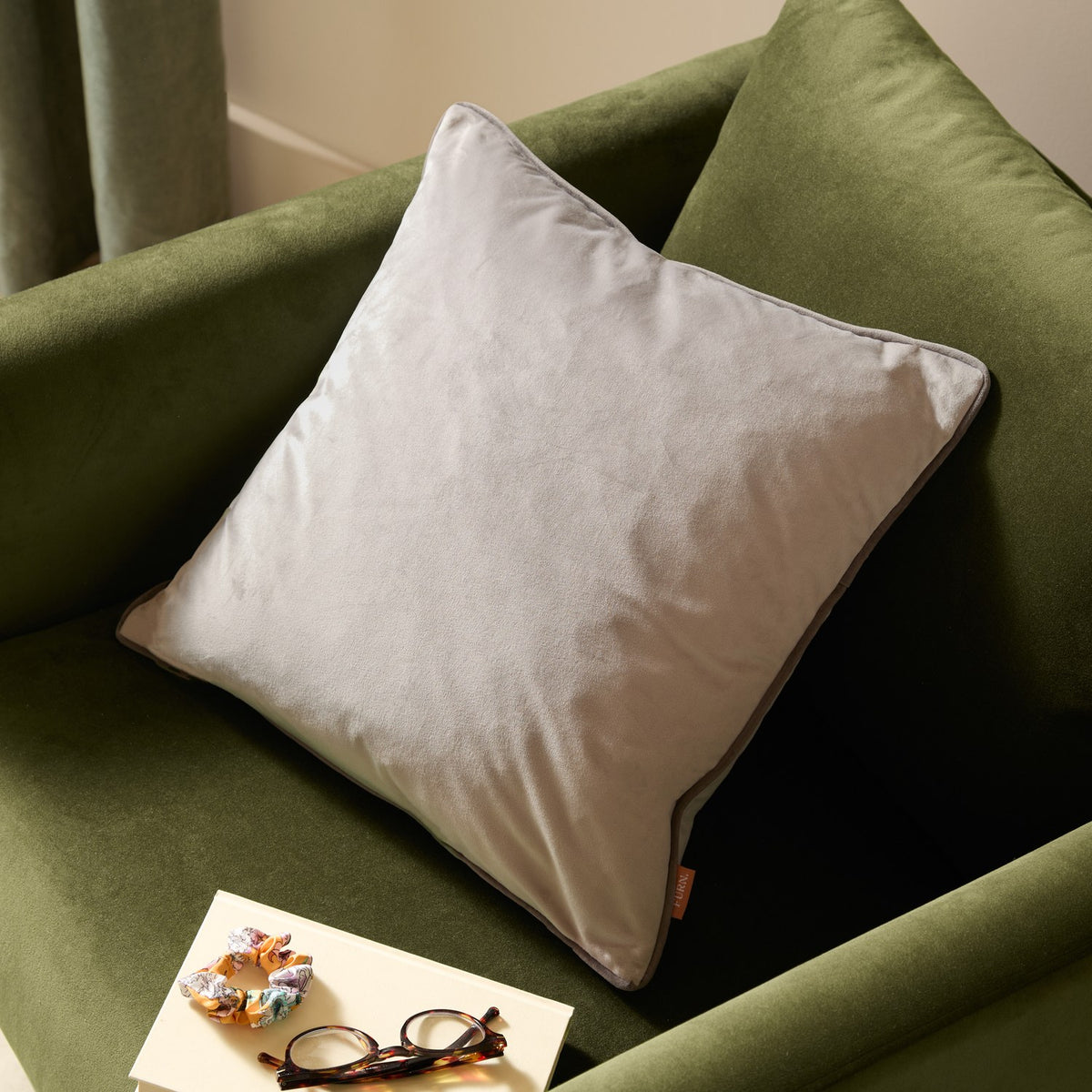 Meridian Plain Velvet Cushion in dove grey with charcoal piped edging, shown on a green velvet armchair beside glasses and a scrunchie.