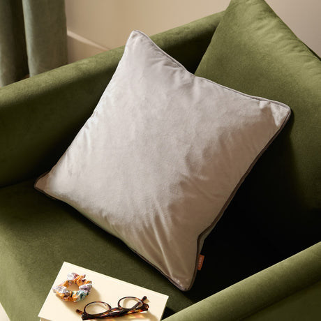 Meridian Plain Velvet Cushion in dove grey with charcoal piped edging, shown on a green velvet armchair beside glasses and a scrunchie.