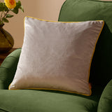 Meridian Plain Velvet Cushion in dove grey with contrasting yellow piped edge, shown on a green armchair beside a vase of yellow flowers.
