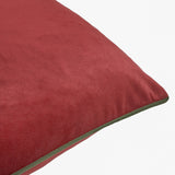 Close-up of the Meridian plain velvet cushion in dark red, showing soft pile fabric and a contrasting fir green piped edge, set against a plain light background.