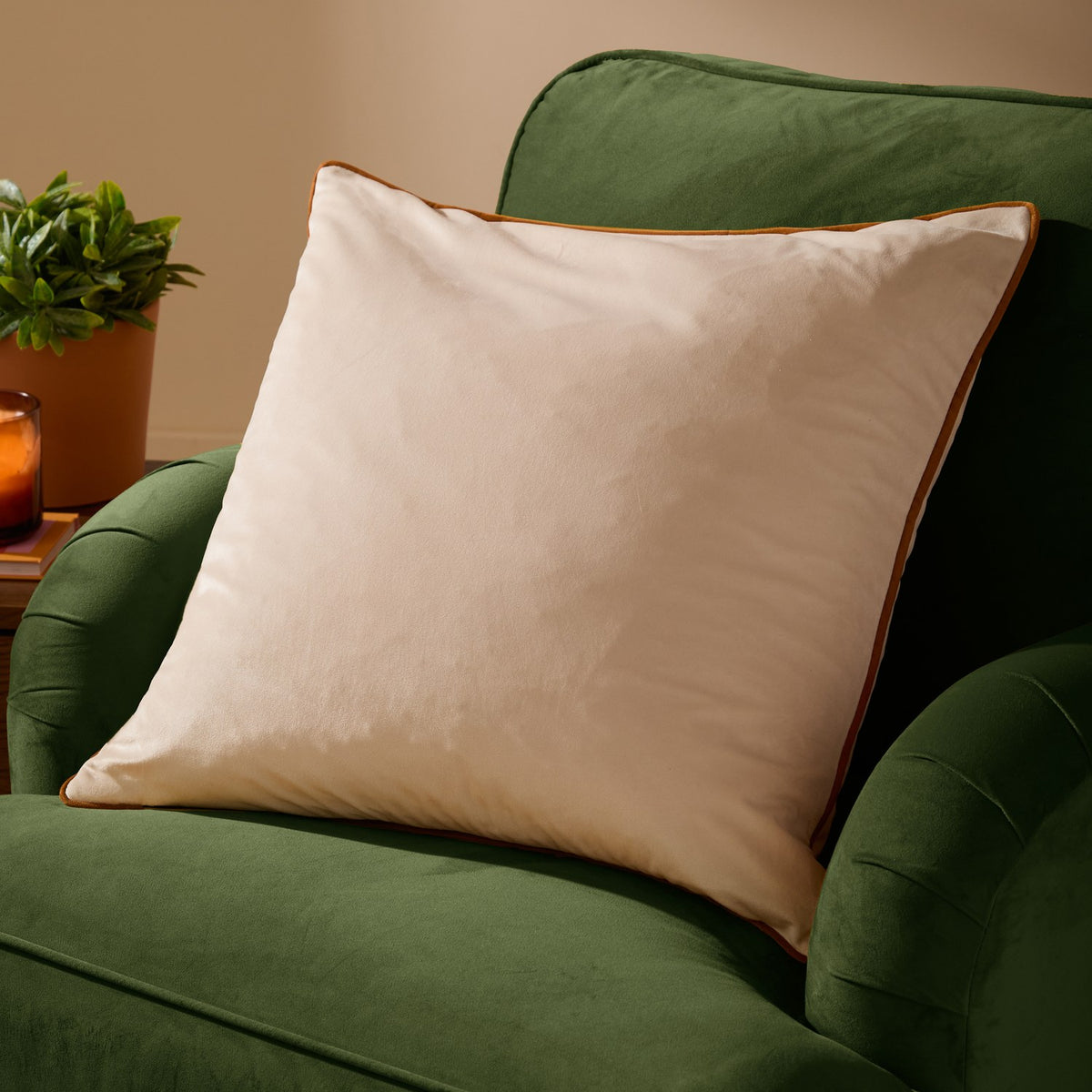 Meridian Plain Velvet Cushion in ecru with contrasting ginger piped edge, shown on a green velvet sofa in a living room setting.