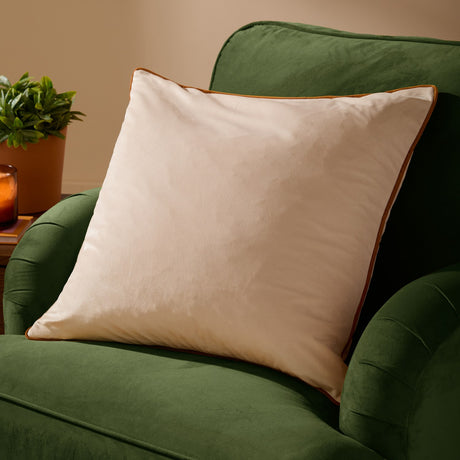 Meridian Plain Velvet Cushion in ecru with contrasting ginger piped edge, shown on a green velvet sofa in a living room setting.
