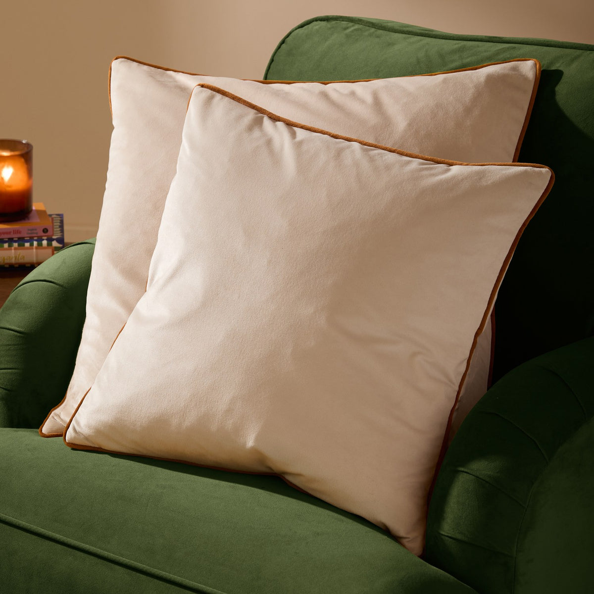 Two Meridian Plain Velvet Cushions in ecru with contrasting ginger piped edges, styled on a green upholstered armchair beside books and a lit candle.