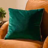 Meridian plain velvet cushion in emerald green with a contrasting blush pink piped edge, shown propped on an orange upholstered armchair.
