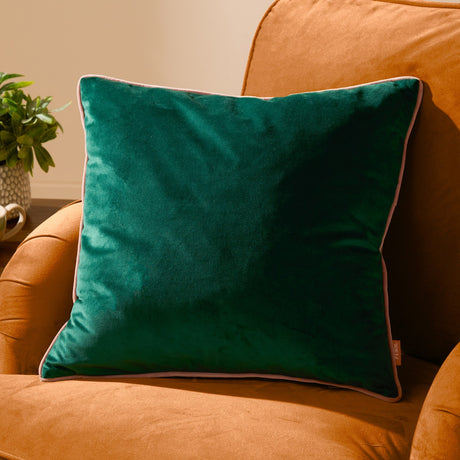 Meridian plain velvet cushion in emerald green with a contrasting blush pink piped edge, shown propped on an orange upholstered armchair.