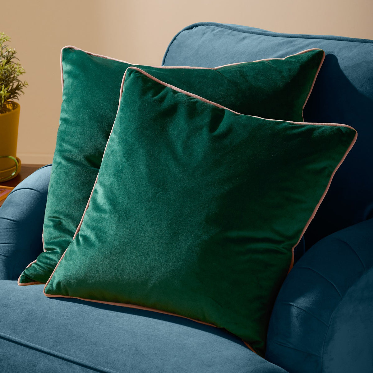 Two Meridian square velvet cushions in emerald green with contrasting blush pink piped edges, styled on a teal upholstered sofa.