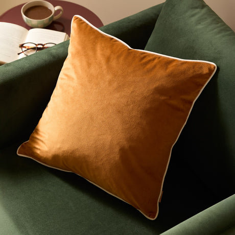 Meridian Plain Velvet Cushion in ginger with ecru piped edging, shown on a green velvet armchair beside a book, glasses and coffee cup.