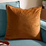 Meridian Plain Velvet Cushion in ginger with olive piped trim, square shape, shown on a teal velvet sofa in a living room setting.