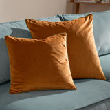 Two square Meridian velvet cushions in ginger with contrasting olive piped trim, styled on a light blue sofa in a living room setting.
