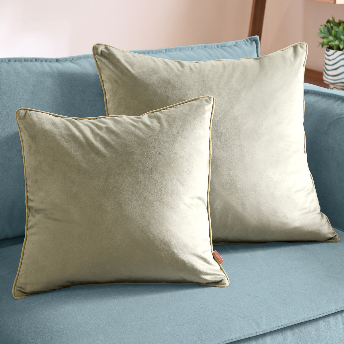 Two Meridian square velvet cushions in sage green with contrasting golden piped trim, styled on a blue sofa.