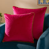Meridian Plain Velvet Cushion in hot pink with contrasting lime green piped edge, shown styled on a teal velvet sofa in a living room setting.