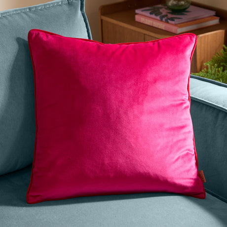 Meridian Plain Velvet Cushion in hot pink with contrasting red piped trim, square scatter size, shown on a light blue sofa beside a wooden side table.